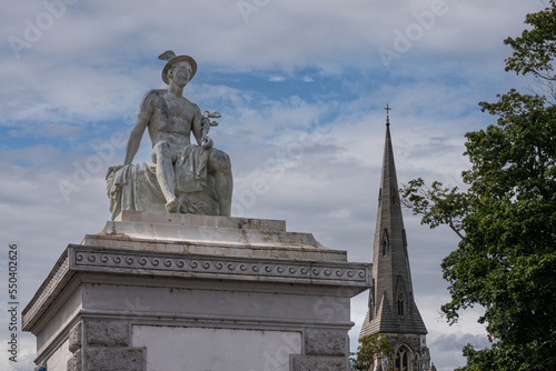 Copenhagen, Denmark - July 24, 2022: Mercurius or Hermes God white stone statue on pillar at east side entrance to Churchill Parken. Blue cloudscape, St. Albans church spire, green foliage photo