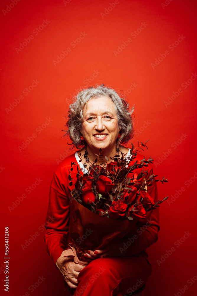 Portrait of a senior woman wearing red clothes, holding a red roses bouquet, over a red background