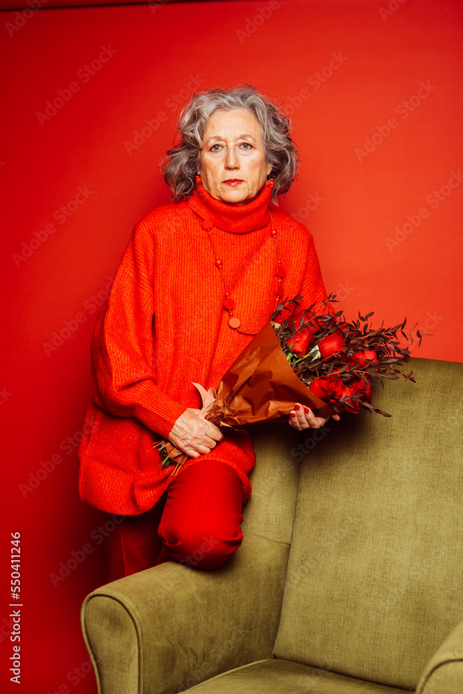 Portrait of a senior woman wearing red clothes and sitting on a green armchair with a bouquet of red roses, over a red background