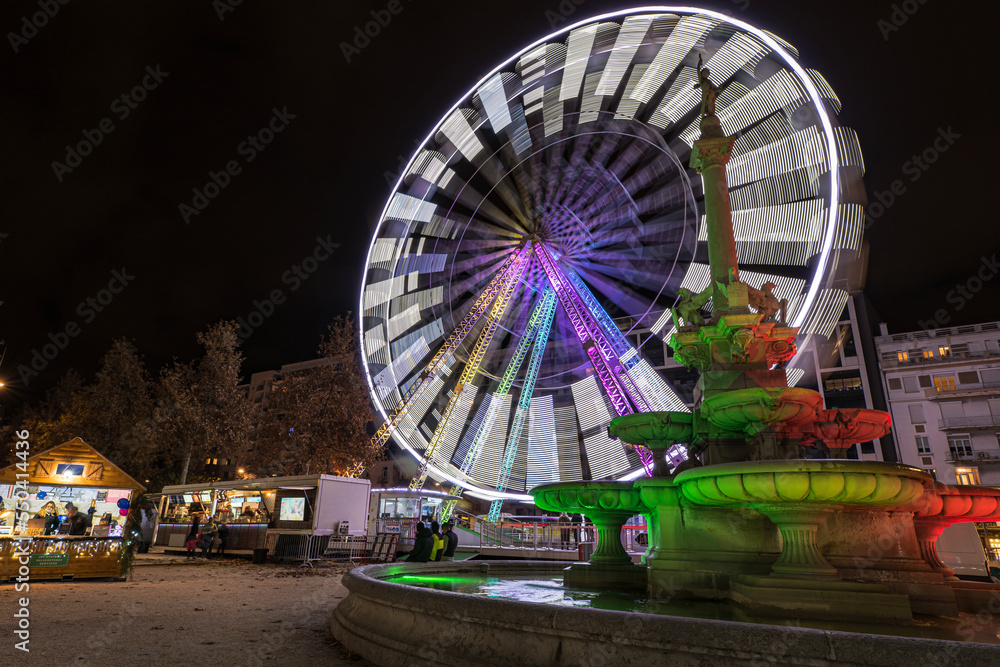Ferris wheel illuminated with lights with stone fountain and Christmas market on a foreground at french Valence city by night