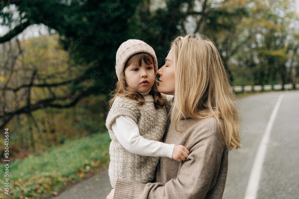 Mom holds her little daughter in her arms and they walk in warm sweaters on an autumn day.