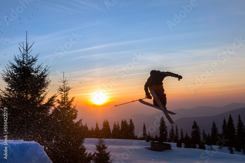 Freestyle skier jumping from kicker in snowy mountains at orange sunset