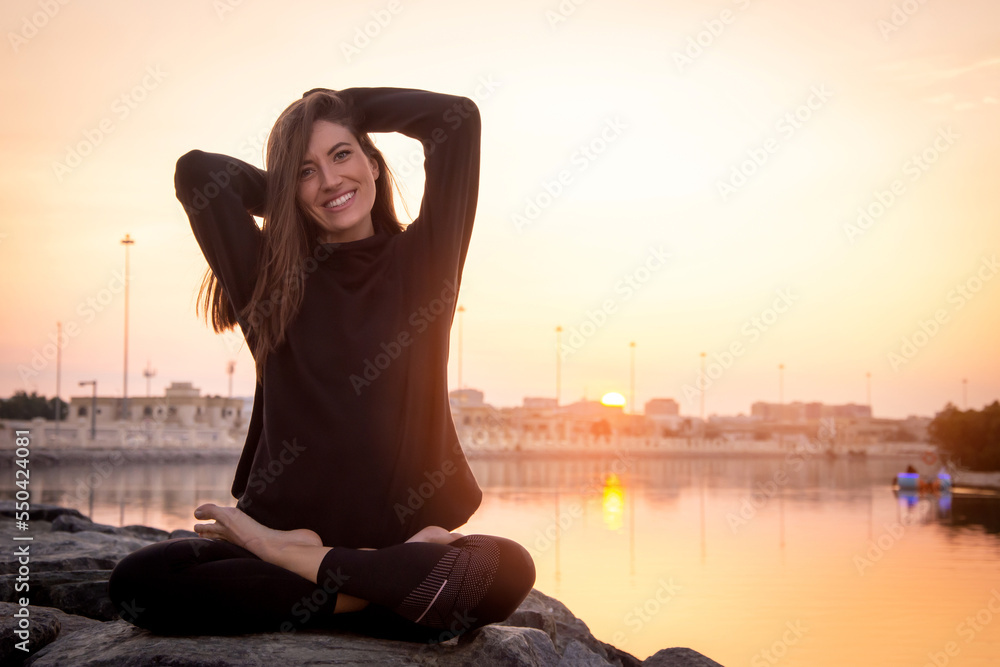 Young beautiful smiling woman sitting with crossed legs preparing for yoga session wearing sportswear iat sunset.