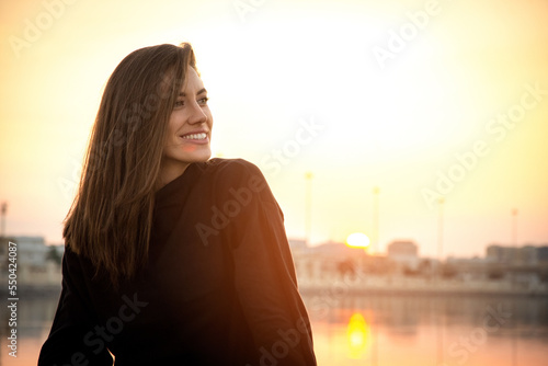 Portrait of young smiling woman sitting outside wearing black sweatshirt at sunset.