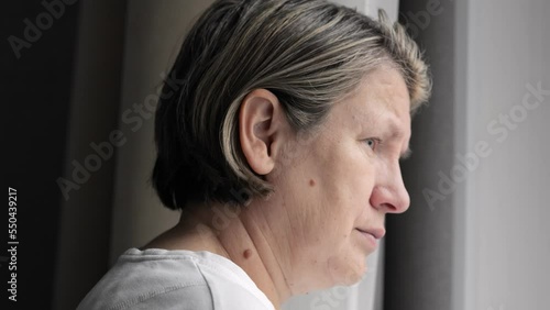 Lonely mature woman stands near window in bedroom looking distracted. Female person feels depressed and indifferent staying at home alone closeup