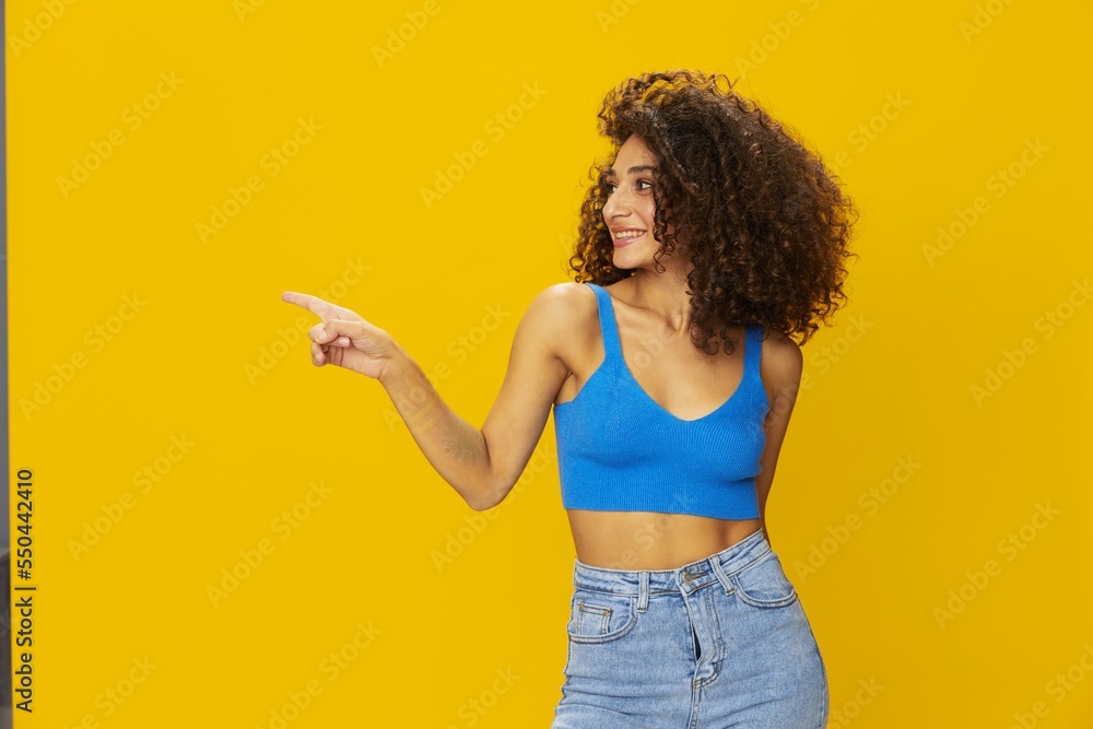 Woman with curly afro hair in a blue t-shirt on. yellow background signs with her hands, look into the camera, smile with teeth and happiness, copy space Woman with curly afro hair in a blue t-shirt on. yellow background signs with her hands, look into the camera, smile with teeth and happiness, copy space