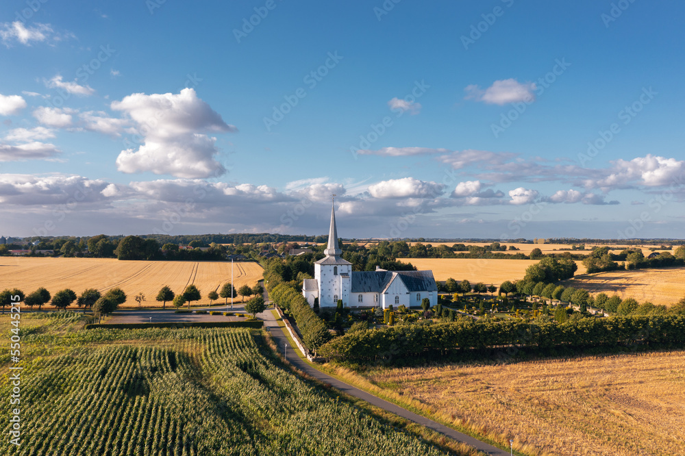 Summer rural scenic landscape in Denmark: traditional Danish white church with a graveyard, surrounded by wheat and corn fields in a countryside