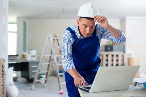 Foreman in overall using laptop in construction site, repair works in apartment.