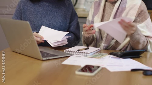 two female in warm clothes sitting kitchen table look through papers bills documents counting calculator expenses payments utility fees taxes. laptop smartphone on table. concept home budget problems photo