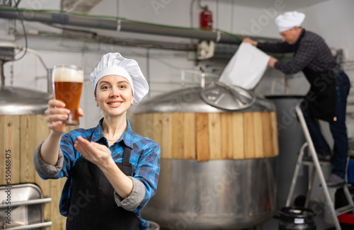 Cheerful young female brewer recommending craft beer, holding out glass with drink while standing against background of fermenters in brewery