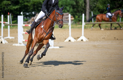 Jumping horse with rider in the course, photographed from the front, another rider out of focus in the background..