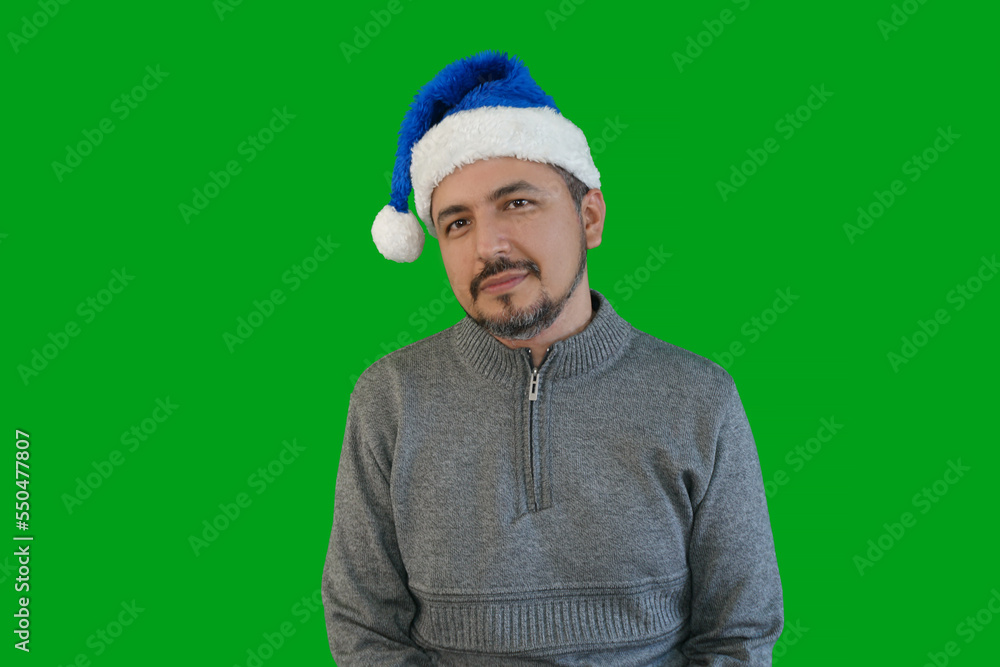 Latino man with a beard, wears a gray sweater, a blue Christmas hat and a green background, he is sitting with a poker face.Chistmas concept. Latino man with a beard, wears a gray sweater, a blue Christmas hat and a green background, he is sitting with a poker face.Chistmas concept.