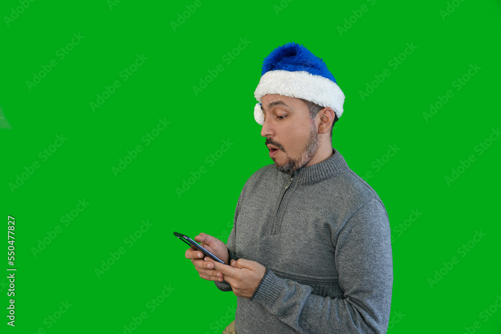 Old Latino man dressed in gray buso and blue Christmas hat, is in profile while staring in amazement at his cell phone screen.Chistmas concept. Old Latino man dressed in gray buso and blue Christmas hat, is in profile while staring in amazement at his cell phone screen.Chistmas concept.