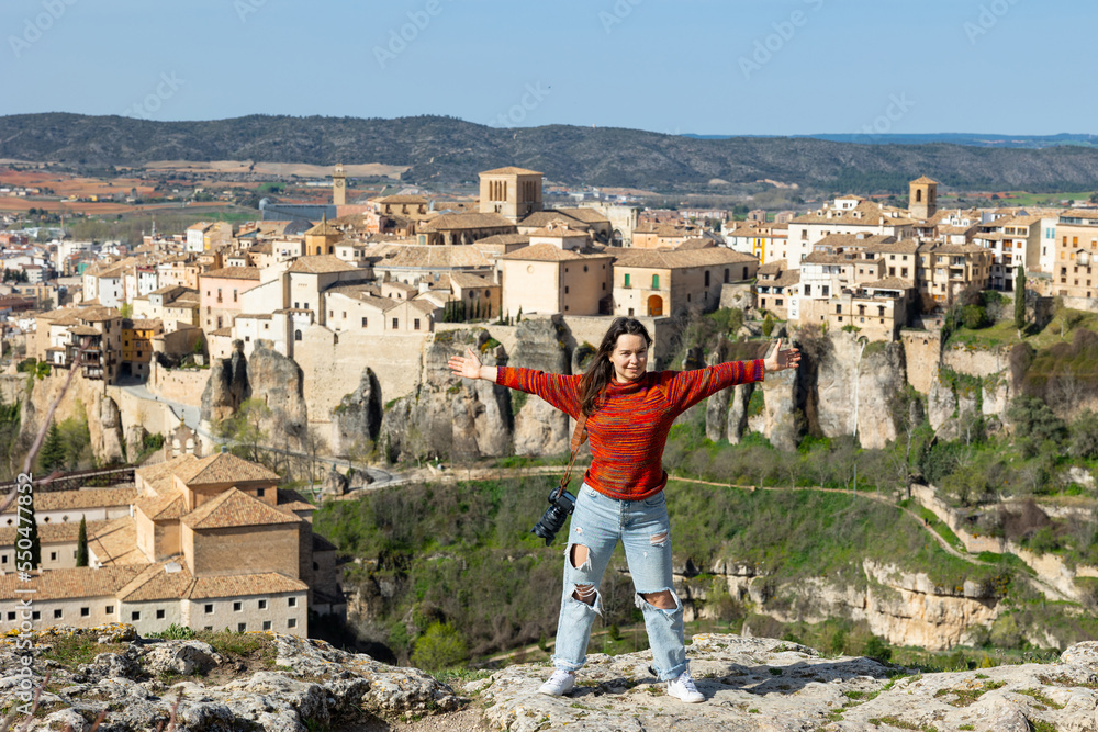Young woman tourist with a camera, who climbed to the top of a cliff against the background of the city of Cuenca, stands ..and enjoys freedom spreading her arms in different directions, Spain