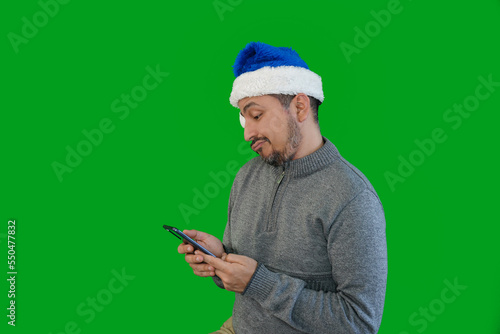 Old Latino man dressed in gray buso and blue Christmas hat, is in profile while looking resignedly at his cell phone screen.Chistmas concept. photo