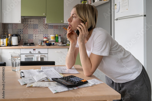 Worried middle-aged European woman housewife managing household finances, making phone call while standing at table with bills and invoices in kitchen. Money management, savings and economy concept photo