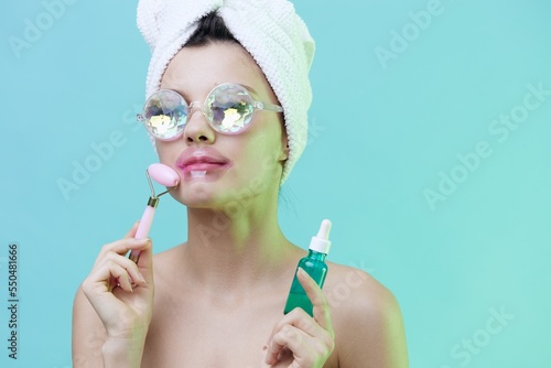  Horizontal photo, a woman with gorgeous skin on a blue background in a towel on her head and body and luxurious glasses and patches on her lips takes care of her face with serum