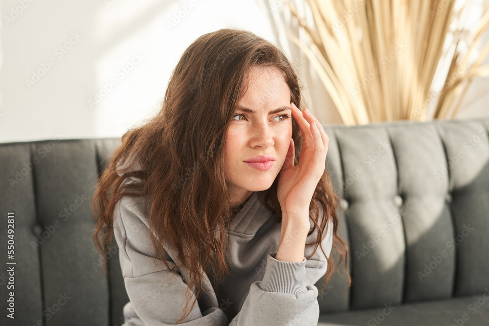 young woman sits on couch discomfort and squints. bored and sad woman with no mood and prop up head with hand young woman sits on couch discomfort and squints. bored and sad woman with no mood and prop up head with hand