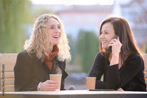 Two happy women friends having fun time together at street cafe laughing and talking on cellphone