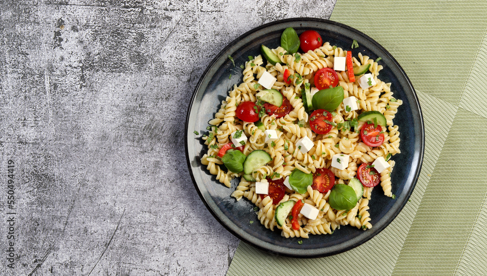Pasta Salad with tomatoes, bell peppers and feta cheese on a round plate on a dark gray background. Top view, flat lay