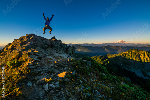 Athletic adventurous male hiker, triumphantly jumping in the air on top of a mountain with Mount Rainier in the background during a beautiful sunrise. 