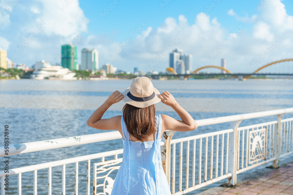 Woman Traveler with blue dress visiting in Da Nang city. Tourist sightseeing the river view with Dragon bridge. Landmark and popular for tourist attraction. Vietnam and Southeast Asia travel concept