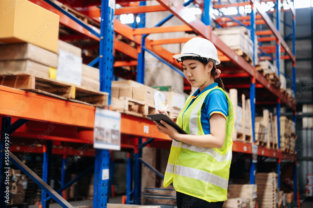 Storage worker in uniform Wearing Hard Hat Checks Stock and Inventory with Digital Tablet Computer in the Retail Warehouse full of Shelves with Goods. Storage worker in uniform Wearing Hard Hat Checks Stock and Inventory with Digital Tablet Computer in the Retail Warehouse full of Shelves with Goods.
