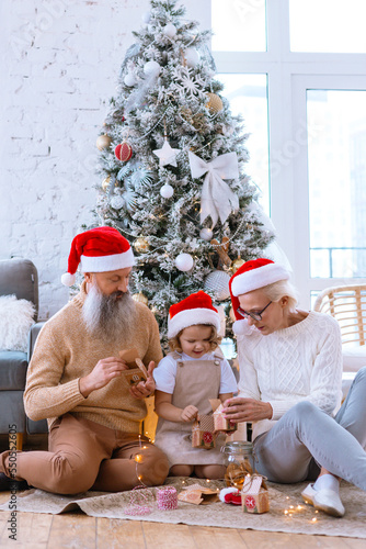 Christmas family grandmother  grandfather and granddaughter sit on sofa near Christmas tree and open advent calendar  pretty girl and active happy seniors give each other gift box