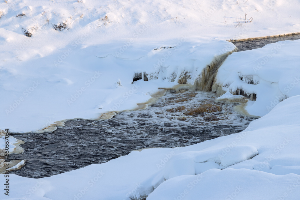 Top view of an icy freezing waterfall. Winter river landscape. Stormy streams of water among ice and snow. Cold winter weather. Tosnensky waterfall, Tosna river, Ulyanovka, Leningrad Oblast, Russia.