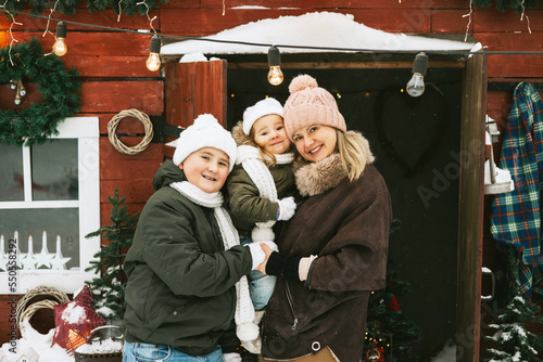 family mother, siblings girl sister and teenage cute boy brother in knitted sweater and hat having fun with first snow at porch of country house, concept of winter spirit and Christmas.