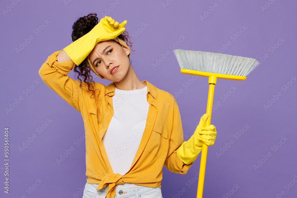 Young sad housekeeper woman wear yellow shirt rubber gloves tidy up hold broom sweeps floor clean house put hand on head forehead isolated on plain pastel purple background studio. Housework concept. Young sad housekeeper woman wear yellow shirt rubber gloves tidy up hold broom sweeps floor clean house put hand on head forehead isolated on plain pastel purple background studio. Housework concept.