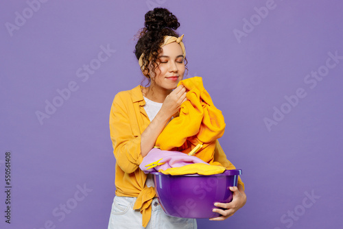 Young housekeeper woman wear yellow shirt tidy up hold basin with clean clothes after laundry sniff scent of washing powder isolated on plain pastel light purple background studio. Housework concept.