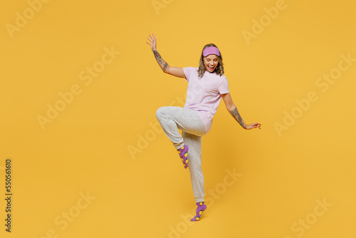 Full body side view young woman she wears purple pyjamas jam sleep eye mask rest relax at home spread hands raise up leg posing isolated on plain yellow background studio portrait. Night nap concept.