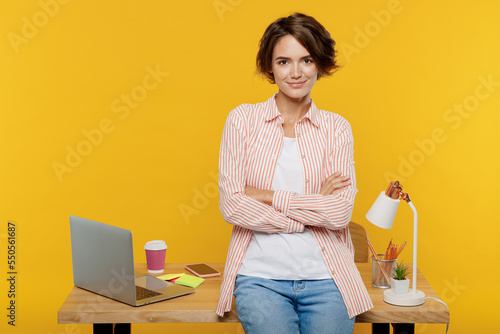 Young confident fun employee business woman wear casual shirt work stand near office desk with pc laptop hold hands crossed folded isolated on plain yellow color background Achievement career concept. photo