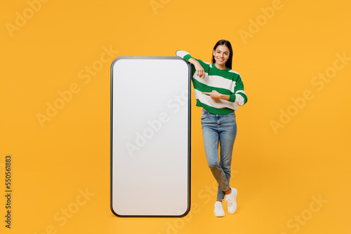 Full body young latin woman wear casual cozy green knitted sweater point index finger big huge blank screen mobile cell phone smartphone with area isolated on plain yellow background studio portrait.
