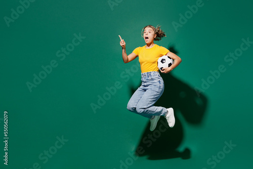 Full body young fun woman fan in basic yellow t-shirt cheer up support football sport team hold in hand soccer ball watch tv live stream jump high point finger aside isolated on dark green background.
