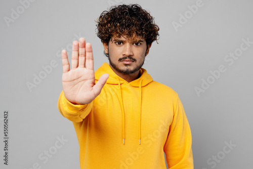 Young strict serious sad indignant Indian man 20s he wear casual yellow hoody showing stop gesture with palm look camera isolated on plain grey background studio portrait. People lifestyle portrait.