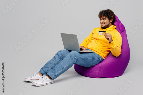 Full body young Indian man 20s he wearing casual yellow hoody using laptop pc computer hold credit bank card doing online shopping order delivery booking tour isolated on plain grey background studio.