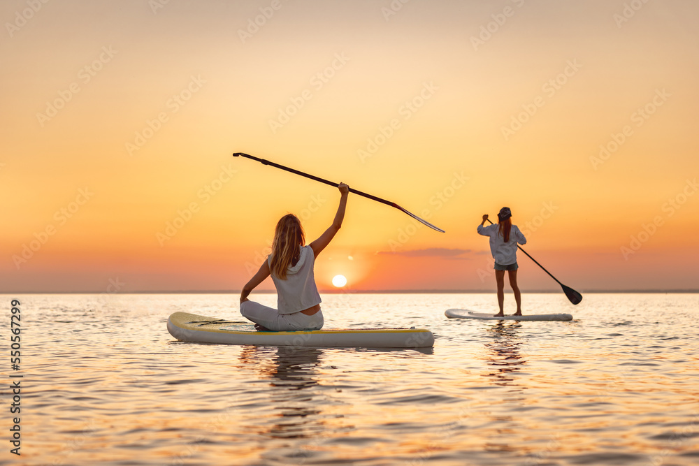 Two happy slim young girls walks at sunset lake on sup stand up paddle boards