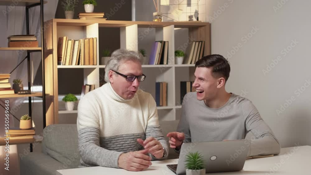 Important information. Happy men. Successful result. Expressive father and son greeting each other with high five getting positive news in laptop in light room interior. Important information. Happy men. Successful result. Expressive father and son greeting each other with high five getting positive news in laptop in light room interior.