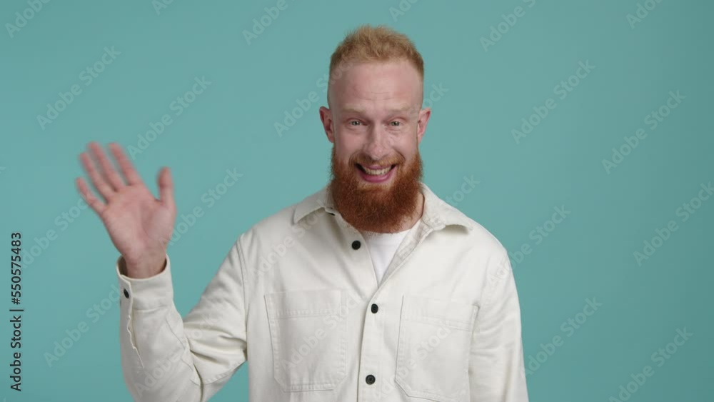 Close-up view of a male model looking positive and cheerful with isolated, blue background behind. Caucasian, red-haired man waving his hand. High quality 4k footage Close-up view of a male model looking positive and cheerful with isolated, blue background behind. Caucasian, red-haired man waving his hand. High quality 4k footage