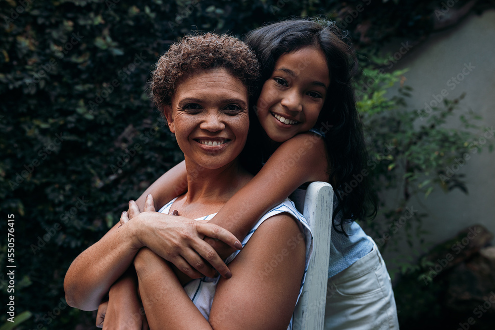 Fototapeta premium Portrait of a girl and her grandmother. Granny sitting on a chair granddaughter hugging her from back.