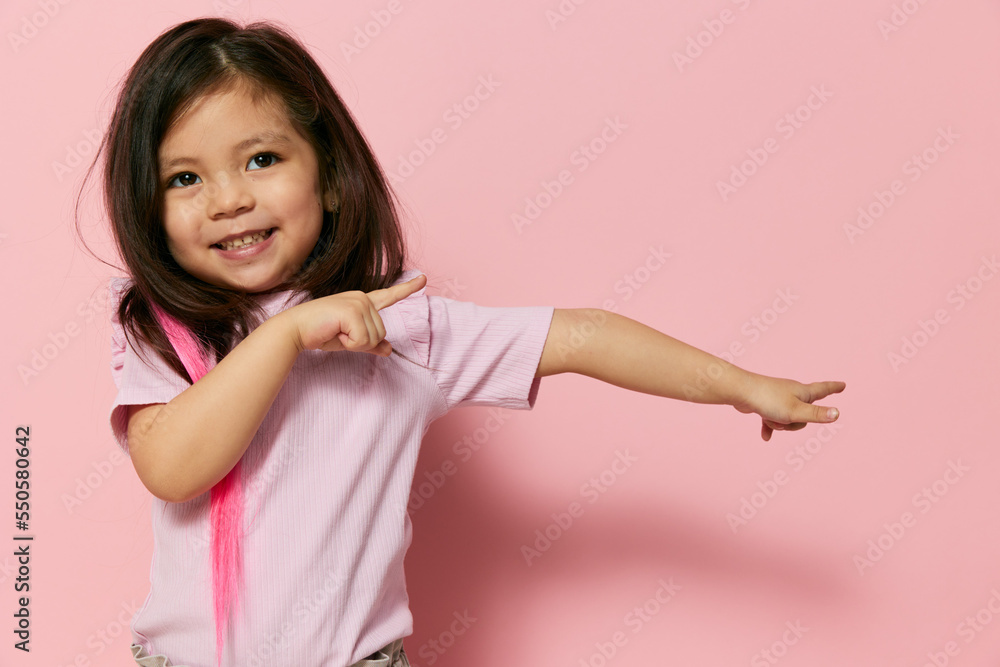 a little beautiful preschool girl stands on a pink background in a pink T-shirt with her hair down playfully looks at the camera and actively points to the side
