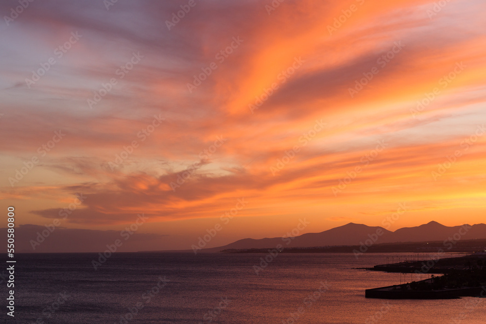 Horizontal seascape of sunset on the beach with silhouettes of mountains in the background and calm sea lapping the coast. Sky with orange, golden and yellow clouds. Lanzarote, Canary Islands, Spain. Horizontal seascape of sunset on the beach with silhouettes of mountains in the background and calm sea lapping the coast. Sky with orange, golden and yellow clouds. Lanzarote, Canary Islands, Spain.