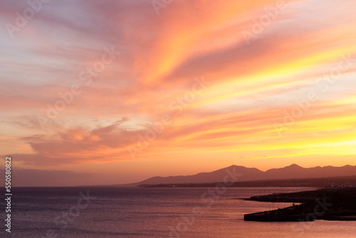 Horizontal seascape of sunset on the beach with silhouettes of mountains in the background and calm sea lapping the coast. Sky with orange, golden and yellow clouds. Lanzarote, Canary Islands, Spain. 