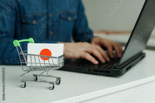 Small shopping cart with flag of Japan on the table. Man using laptop for shopping online.