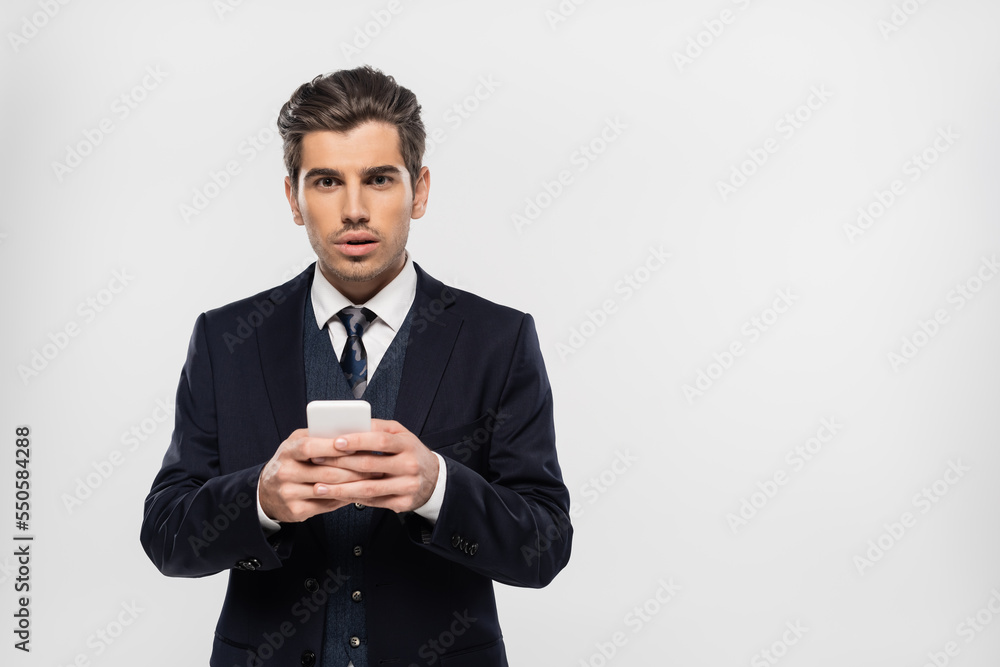 stunned and young businessman in suit messaging on smartphone isolated on grey stunned and young businessman in suit messaging on smartphone isolated on grey