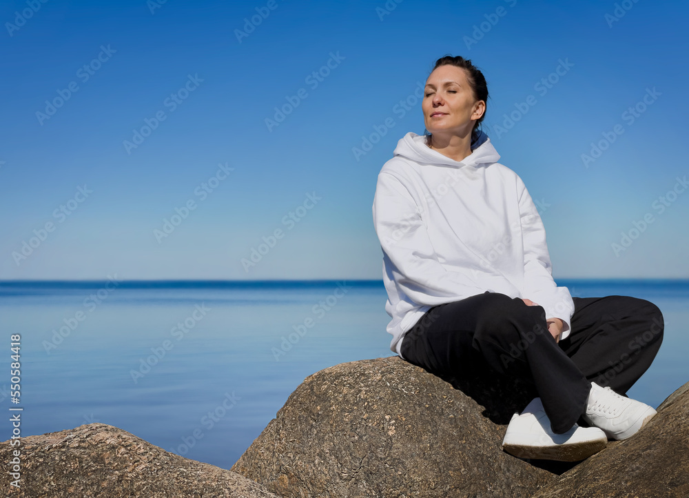 A brunette woman, relaxed, in loose, comfortable clothes, sitting on a rock by the sea, her head turned to the sun, her eyes closed. A brunette woman, relaxed, in loose, comfortable clothes, sitting on a rock by the sea, her head turned to the sun, her eyes closed.