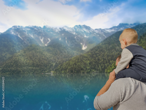 A man with a small child stand on the shore of a mountain lake and look at the mountains. Dad and son. The child is sitting on Dad's shoulders. Family. View from the back. Summer, autumn day.