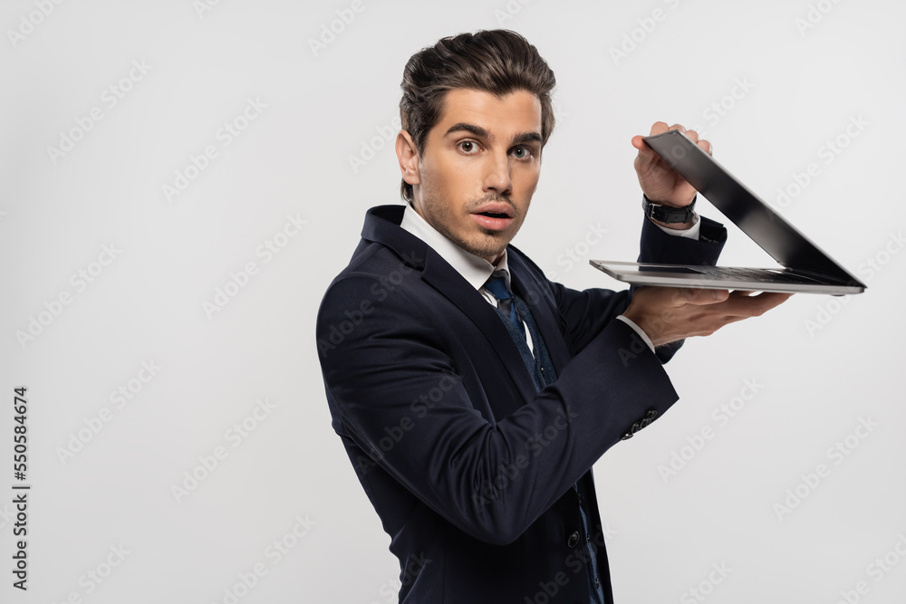 stunned businessman in suit looking at camera and holding laptop isolated on grey stunned businessman in suit looking at camera and holding laptop isolated on grey
